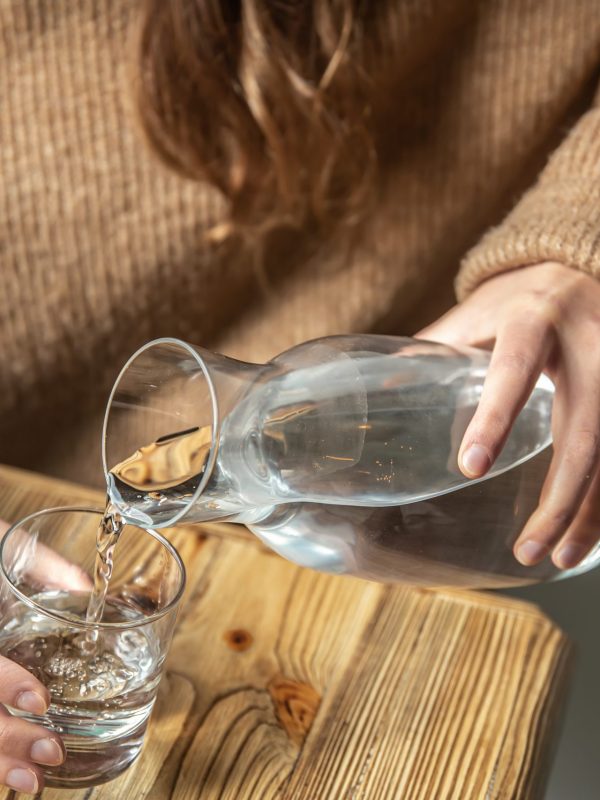 A woman in a cozy sweater pours water into a glass from a glass decanter, the concept of maintaining water balance.